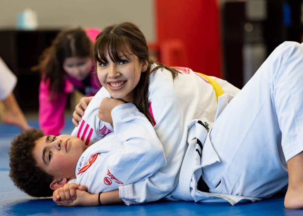 A teen boy and a girl practicing BJJ at Gracie Barra Salt Lake City. The girl is looking at the camera smiling.