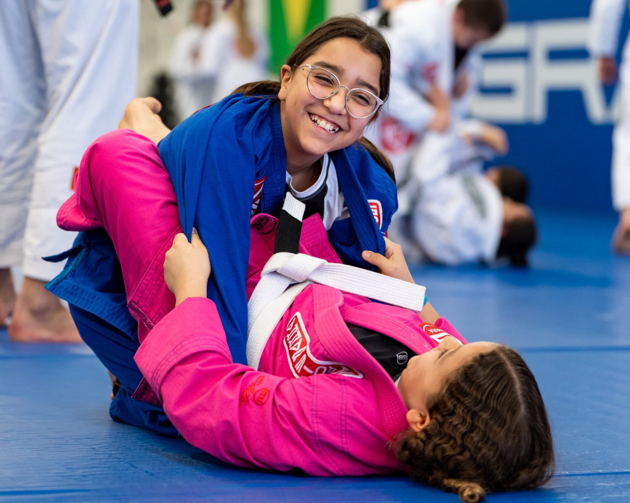 Two girls practicing BJJ, one is wearing prescription glasses and is smiling at the camera, while the other is on the floor.